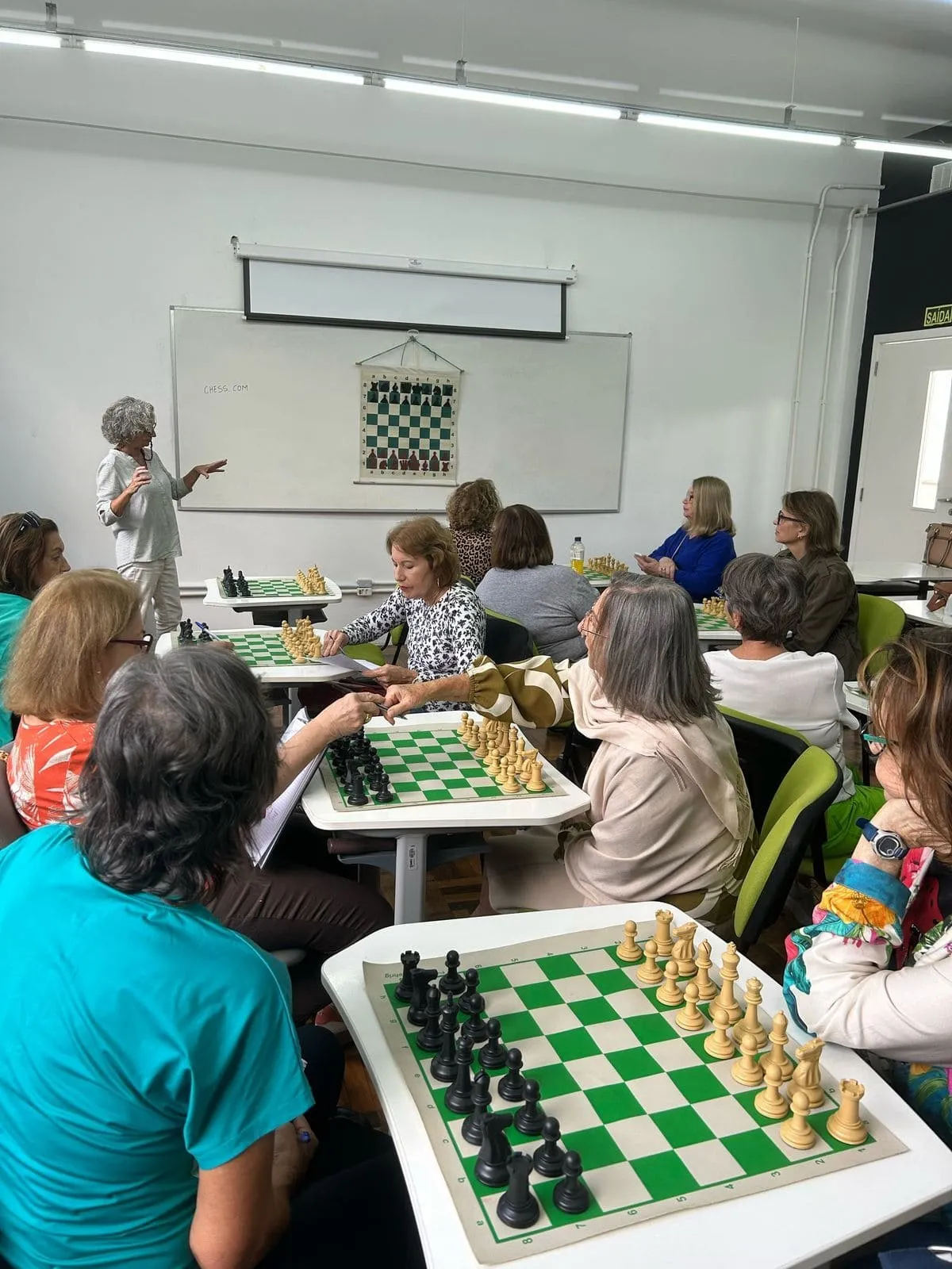 Alunos jogando xadrez durante uma aula livre, com a professora ao fundo